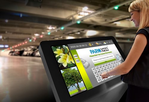 A blonde woman stands at a digital kiosk inside a parking garage. On the touchscreen displaying the Parkxper logo and keybaord she enters a license plate number.