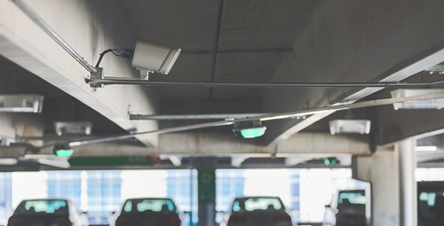 Camera and green parking guidance light suspended from the ceiling of a parking garage with a row of parked cars in the background